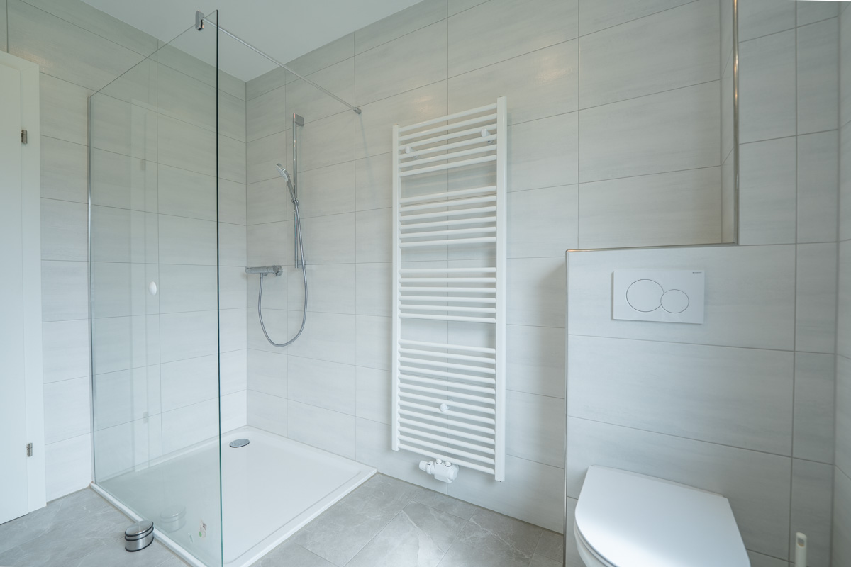 Modern saniertes Badezimmer mit bodengleicher Dusche, Glasduschwand, Handtuchheizkörper und hochwertiger Fliesenoptik.